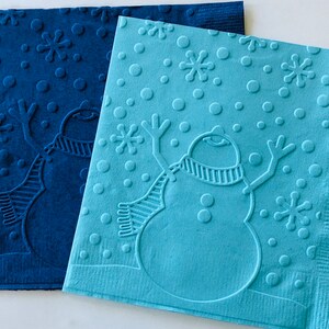 May include: Two embossed paper napkins with a winter theme. One is a deep blue, the other is light blue. Each napkin features a snowman with a scarf, snowflakes, and dots. The napkins are likely for a holiday or winter event.