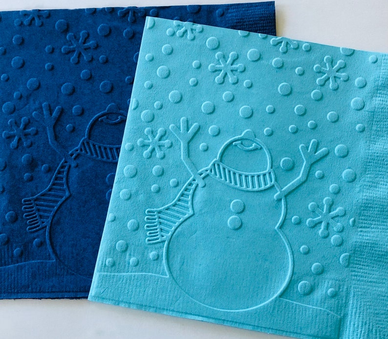 May include: Two embossed paper napkins with a winter theme. One is a deep blue, the other is light blue. Each napkin features a snowman with a scarf, snowflakes, and dots. The napkins are likely for a holiday or winter event.