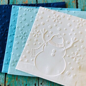 May include: A stack of four square paper napkins in shades of blue. The napkins feature embossed designs of snowflakes, circles, and a snowman with a scarf. The napkins are arranged on a weathered, turquoise wooden surface.