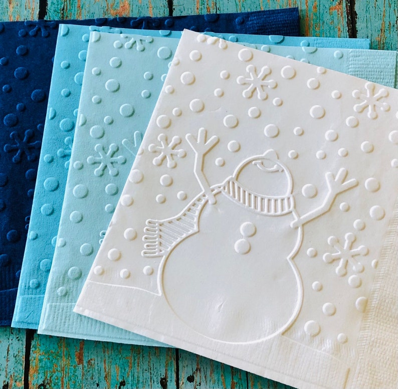 May include: A stack of four square paper napkins in shades of blue. The napkins feature embossed designs of snowflakes, circles, and a snowman with a scarf. The napkins are arranged on a weathered, turquoise wooden surface.