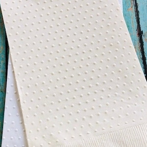 May include: Two off-white paper napkins with a textured, dotted pattern. The bottom edge of each napkin has a ribbed texture. The napkins are stacked on top of each other, with the top napkin slightly angled.
