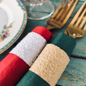 May include: Two gold-toned metal forks wrapped in napkins. One fork is wrapped in a red napkin with a white floral-patterned paper band. The other fork is wrapped in a dark green napkin with a light beige floral-patterned paper band.