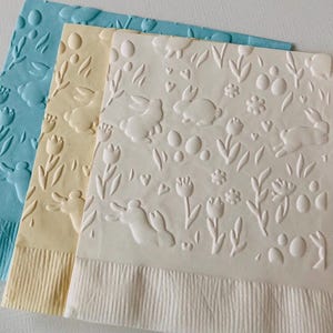 May include: Three decorative paper napkins in pastel colors: light blue, pale yellow, and white. Each napkin features an embossed design of Easter-themed elements, including bunnies, eggs, flowers, and hearts. The bottom edge of each napkin has a textured border.