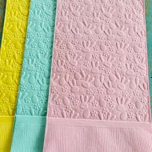 May include: Three Easter-themed towels in yellow, teal, and pink. Each towel features a repeating embossed pattern of bunnies, eggs, and carrots. The bottom of each towel has a textured, ribbed border.