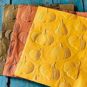 May include: Three fall-themed napkins in shades of green, orange, and yellow. Each napkin features an embossed pumpkin design. The napkins are arranged on a weathered blue wooden surface, creating a rustic autumn aesthetic.