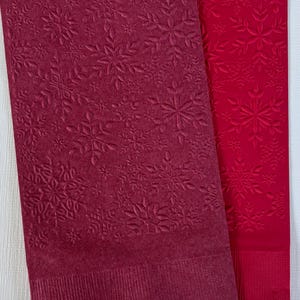 May include: Two rectangular paper napkins. One is a deep burgundy colour, and the other is a bright red. Both napkins feature an embossed snowflake pattern. The burgundy napkin has a more detailed snowflake design, while the red napkin has a simpler pattern.