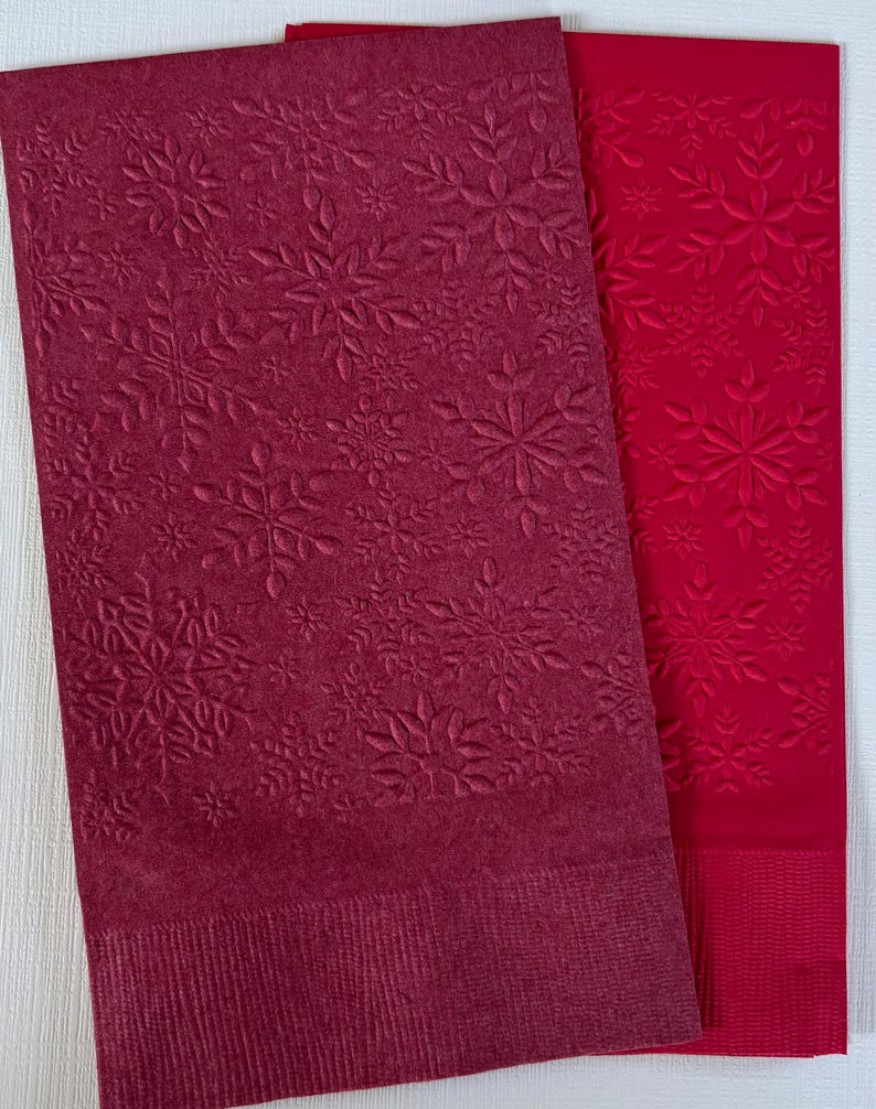 May include: Two rectangular paper napkins. One is a deep burgundy colour, and the other is a bright red. Both napkins feature an embossed snowflake pattern. The burgundy napkin has a more detailed snowflake design, while the red napkin has a simpler pattern.