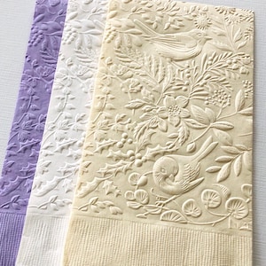 May include: Three decorative paper napkins with embossed floral and bird designs. The napkins are in shades of lavender, white, and cream. The bottom edge of each napkin has a textured border.