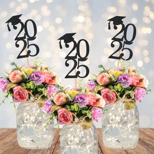 May include: Three glass jars with fairy lights, each holding a floral arrangement of pink, purple, and yellow roses. Black glitter graduation cap toppers with the year 2025 are placed in the center of each arrangement. The jars are tied with twine.