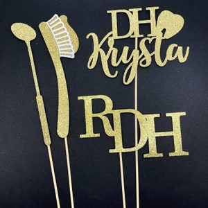 May include: Gold glitter cake toppers with the text "DH Krysta RDH" and a small heart. The toppers are on wooden sticks and are perfect for a dental hygienist's graduation party.