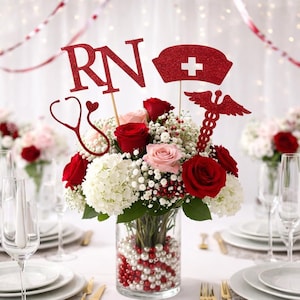 May include: A floral centerpiece in a clear vase, featuring red and pink roses, white hydrangeas, and baby's breath. Embellished with red glitter cutouts spelling "RN", a nurse's cap, and a caduceus symbol. The vase is filled with red and white pearls.