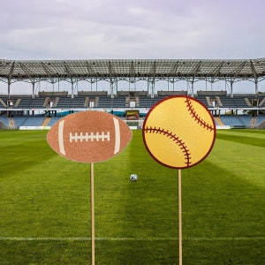 May include: Two glitter-covered sports ball cutouts on wooden sticks. A brown football cutout and a yellow baseball cutout. The cutouts are in front of a green grassy field with a stadium in the background.