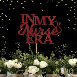 May include: Red glitter cake topper with the text "In My Nurse Era" and a small stethoscope symbol. The topper is on a stick and is placed in a bouquet of white flowers.