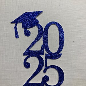 May include: A blue glitter graduation cap and the numbers 2025. The cap is on top of the number 2, and the numbers are vertically aligned. The background is a textured white surface. The image is suitable for graduation announcements or decorations.