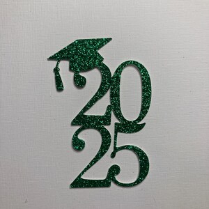 May include: A green glitter graduation cap and the numbers "2025" cut out of green glitter material. The graduation cap is positioned above the numbers, with a tassel hanging down. The background is a textured white surface.