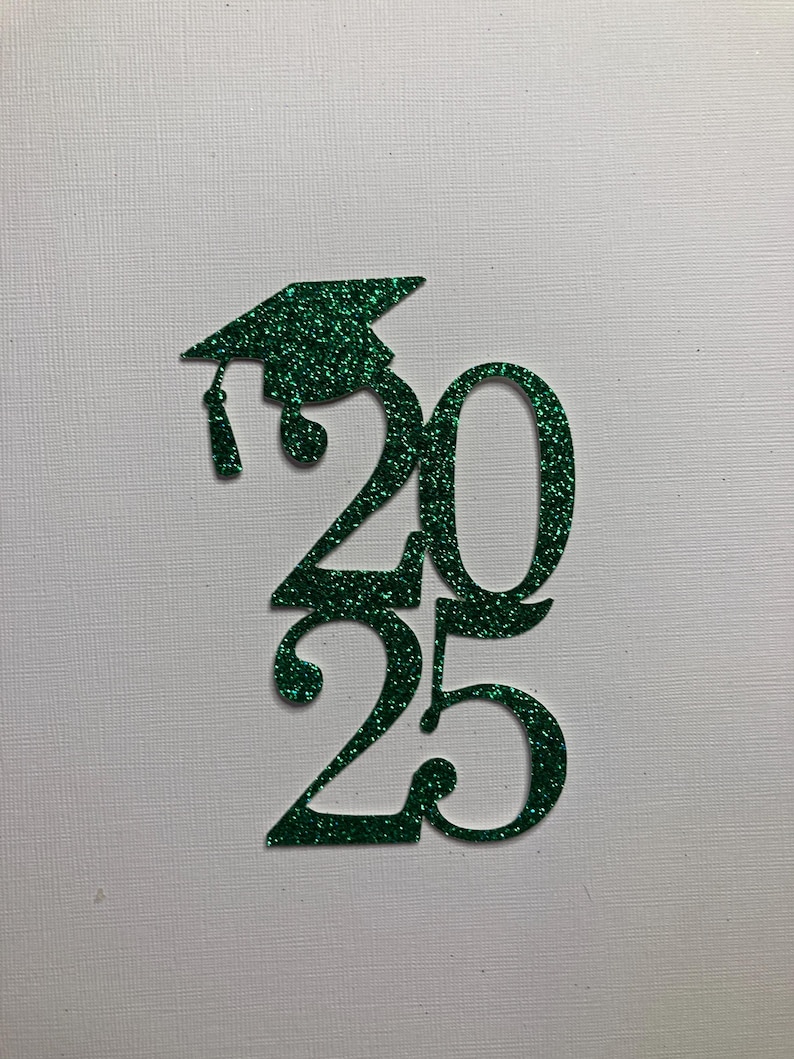 May include: A green glitter graduation cap and the numbers "2025" cut out of green glitter material. The graduation cap is positioned above the numbers, with a tassel hanging down. The background is a textured white surface.