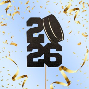 May include: A black glittery cake topper with the number 26 and a hockey puck design. The topper is on a wooden stick, with gold confetti and ribbons against a light blue background.