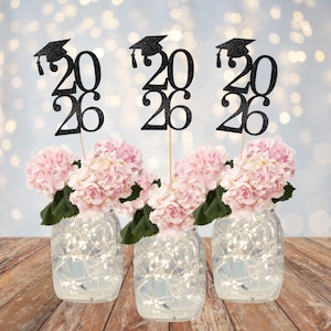 May include: Three mason jars filled with fairy lights and pink hydrangeas. Each jar holds a graduation centerpiece with the year "2026" in black glitter, topped with a graduation cap. The jars sit on a wooden surface.