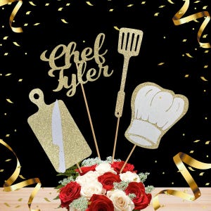 May include: Gold glitter 'Chef Tyler' cake topper featuring a chef's hat, a chopping board, and a spatula. The topper is displayed on a bouquet of red and white roses.