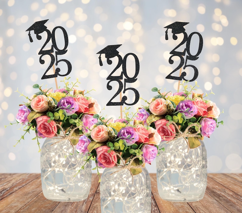 May include: Three floral arrangements in glass jars with fairy lights. Each jar has a bouquet of pink, purple, and yellow flowers. Black glitter graduation cap toppers with the year 2025 are in each arrangement.