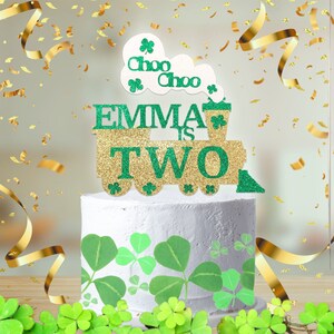 Puede incluir: Un adorno para pastel de purpurina verde y dorada con forma de tren con las palabras "Emma is Two" y "Choo Choo" en él. El tren tiene diseños de trébol.