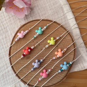 Könnte beinhalten: Sieben Armbänder mit Perlen und Mickey Mouse-förmigen Anhängern in verschiedenen Farben, darunter Rosa, Grün und Gelb. Die Armbänder sind auf weißen Kordeln aufgereiht und auf einer Holzscheibe angeordnet, mit einem neutralfarbenen Stoff im Hintergrund.