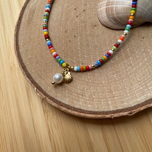 Könnte beinhalten: Eine bunte Perlenkette mit einem goldenen Muschel-Anhänger und einer weißen Perle. Die Kette liegt auf einer Holzoberfläche.