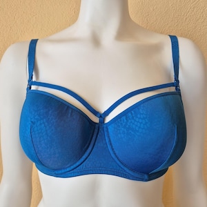 May include: A blue bra with a textured pattern. The bra has adjustable straps and a unique design with additional straps across the upper cups. The bra is displayed on a white mannequin.