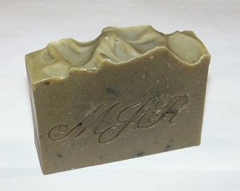 May include: A rectangular bar of handmade soap with a textured, wavy top. The soap is a muted green color with darker flecks and the carved initials "MJR" on the front. The soap appears to be unscented.