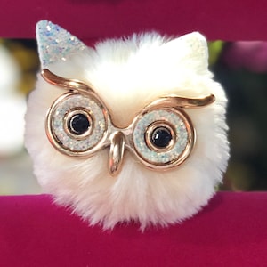 May include: A white fluffy owl charm with gold glitter eyes and a gold glitter headband. The owl is sitting on a pink surface.