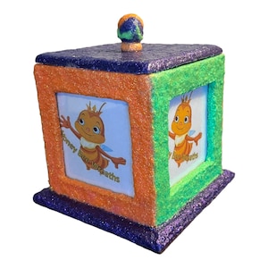 May include: A small, square, orange, green, and purple glitter box with a bee image on each side. The box has a purple lid with a small, orange, green, and purple glitter knob. The bee image on the front of the box says "Honey Bee Wreaths".
