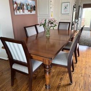 May include: A wooden dining table with a rich, dark finish, surrounded by six chairs with light-colored upholstered seats and striped backs. The table is set on a hardwood floor, with a painting on the wall in the background.