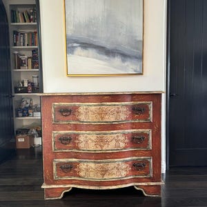 May include: A three-drawer chest with a curved front and painted floral designs. The chest is painted in shades of red, white, and cream. The drawers have antique-style metal handles.