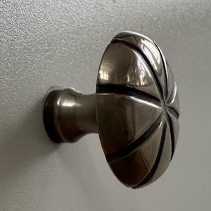 May include: A silver-toned cabinet knob with a decorative, starburst-like design on the rounded face. The knob is mounted on a light gray surface, showcasing its metallic sheen and detailed pattern.