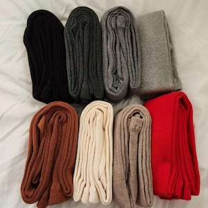 May include: A collection of eight pairs of folded leggings arranged in two rows. The colors include black, dark gray, medium gray, light gray, rust brown, cream, beige, and bright red. Each pair features a ribbed knit texture throughout.
