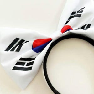 May include: White bow headband featuring the South Korean flag design. The bow has black, red, and blue accents. The headband is black.