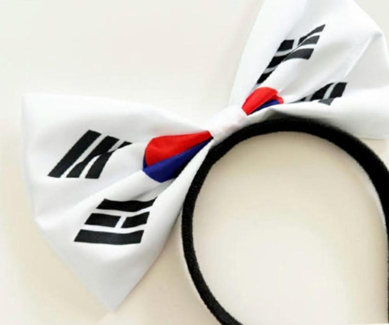 May include: White bow headband featuring the South Korean flag design. The bow has black, red, and blue accents. The headband is black.