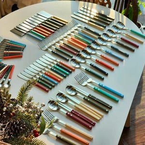 Puede incluir: Un juego de cubiertos coloridos con mangos de madera. El juego incluye cuchillos, tenedores, cucharas y palillos. Los cubiertos están dispuestos sobre una mesa blanca.