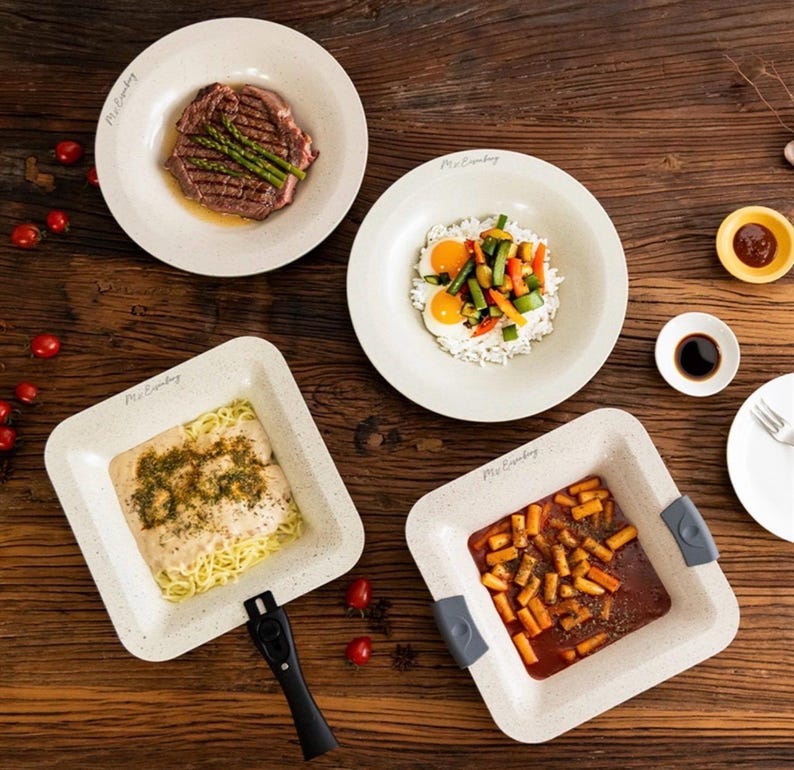 Puede incluir: Vista a&eacute;rea de una mesa de madera con cuatro platos de comida. Los platos beige llevan el texto "Mr. Eatsby". La comida incluye bistec con esp&aacute;rragos, arroz con verduras y huevos, pasta y un plato con salsa roja y trozos de naranja.