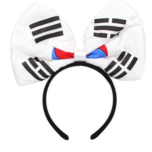 May include: A white headband with a large bow featuring the South Korean flag design. The bow has black stripes and the Taegeuk symbol in red and blue. The headband itself is black.