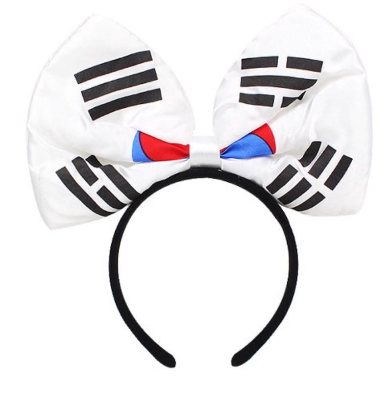 May include: A white headband with a large bow featuring the South Korean flag design. The bow has black stripes and the Taegeuk symbol in red and blue. The headband itself is black.