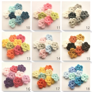 Op de afbeelding: Assortiment van gehaakte bloemenversieringen in verschillende kleuren, waaronder blauw, roze, geel, groen, oranje, paars en wit. De bloemen hebben een diameter van ongeveer 2,5 cm.
