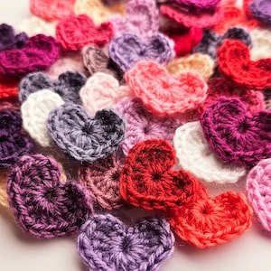 Peut inclure: Une collection de cœurs en crochet de différentes couleurs, notamment rouge, rose, violet et blanc. Les cœurs sont disposés de manière aléatoire, créant un affichage visuellement attrayant et texturé.
