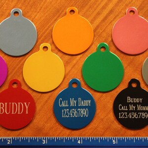 May include: A collection of colorful, round pet ID tags in various colors, including red, blue, and black. Some tags have engraved text such as "Buddy" and contact information. The tags are made of metal.