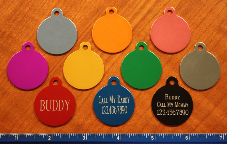 May include: A collection of colorful, round pet ID tags in various colors, including red, blue, and black. Some tags have engraved text such as "Buddy" and contact information. The tags are made of metal.