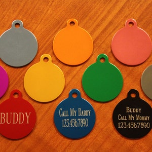 May include: A collection of ten colorful, round pet ID tags. The tags are in various colors, including gray, orange, pink, purple, yellow, green, and blue. Some tags have engraved text, such as "Buddy" and phone numbers.