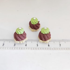 May include: Three miniature desserts with a light brown base, dark red swirl, and green frosting topped with a silver bead. The desserts are arranged on a white surface next to a ruler.