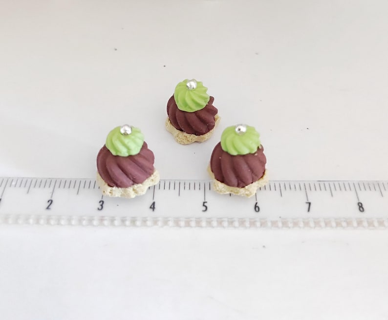 May include: Three miniature desserts with a light brown base, dark red swirl, and green frosting topped with a silver bead. The desserts are arranged on a white surface next to a ruler.