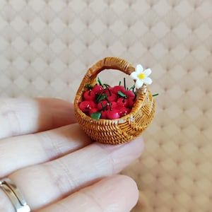 May include: Miniature woven basket filled with red cherries and green leaves. A small white flower with a yellow center adorns the basket's rim. The basket is held in a hand, showcasing the small size of the fruit basket.