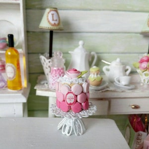 Peut inclure: Un gâteau miniature rose et blanc décoré de macarons et d'une fleur sur un support à gâteau blanc. Le gâteau est sur une table blanche avec un fond blanc et vert.
